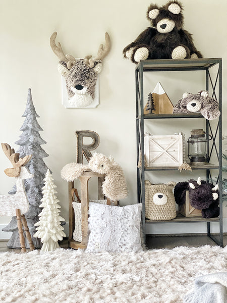 A plush deer wall mount in a woodland themed nursery. The stuffed deer head is made with soft brown and white fabric with crocheted ears. Other forest decor items include three tree sculptures and a brown bear stuffed animal.