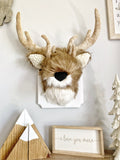 A plush deer head made by ClaraLoo mounted on a wooden plaque in a woodland themed bedroom. The stuffed deer is made with soft brown and white faux fur with crocheted ears. The wooden plaque is painted white.
