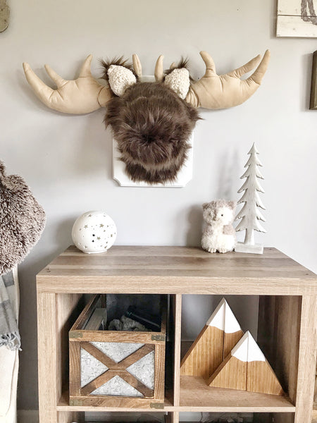 A plush moose head mounted on a white wooden plaque in a woodland themed nursery. This plush moose wall mount is handmade by ClaraLoo with soft brown faux fur and crocheted ears.